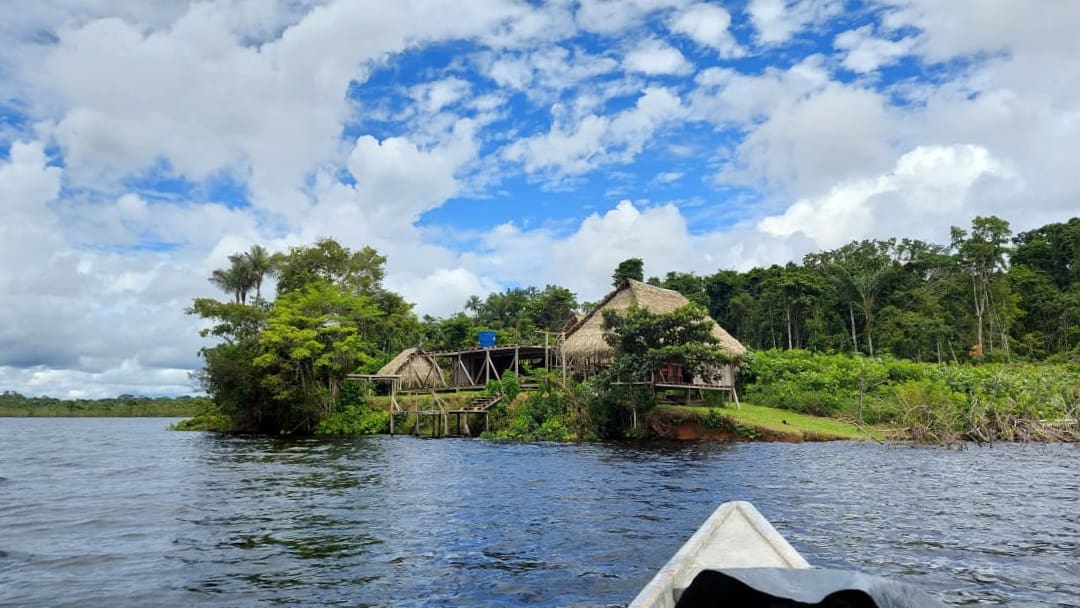 looking over the front of a canoe, we can see a few huts built on the grassy shore with a forest in the background, blue sky, and puffy white clouds