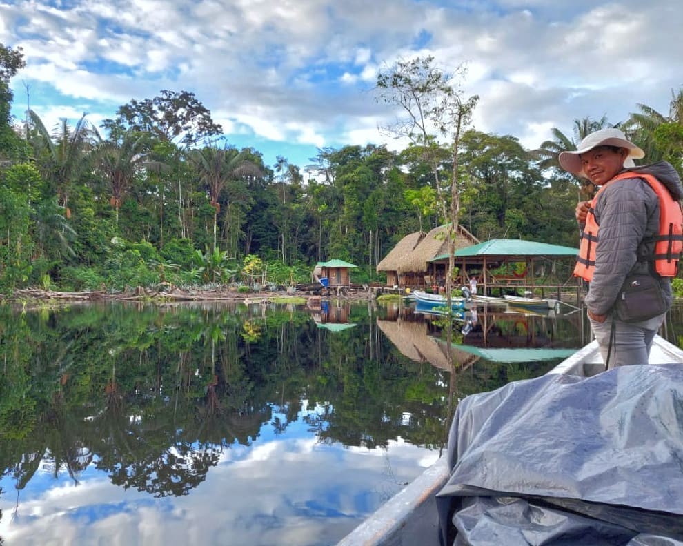 A man stands in the front of a canoe arriving at a pier near a small community in the rainforest